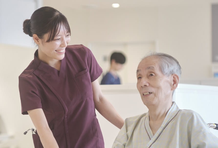 車椅子の高齢の患者さまに寄り添い笑顔で語りかける看護師